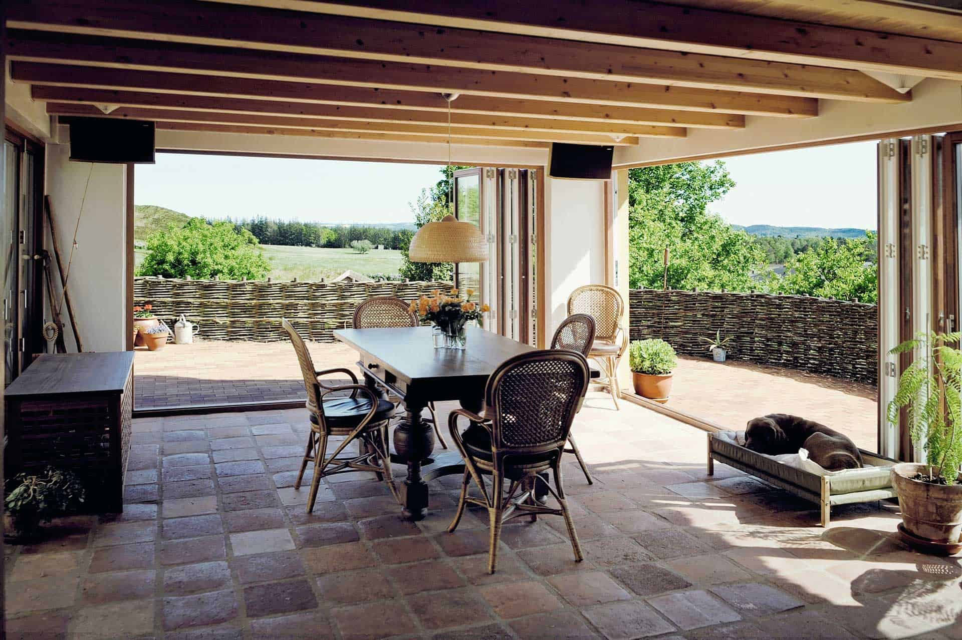 Lyst indendørs spiseområde med store vinduer der vender ud mod en grøn have og klar himmel.