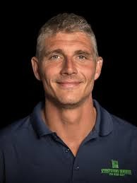 Højde- og breddesiddende mand med smilende ansigt i mørk baggrund.