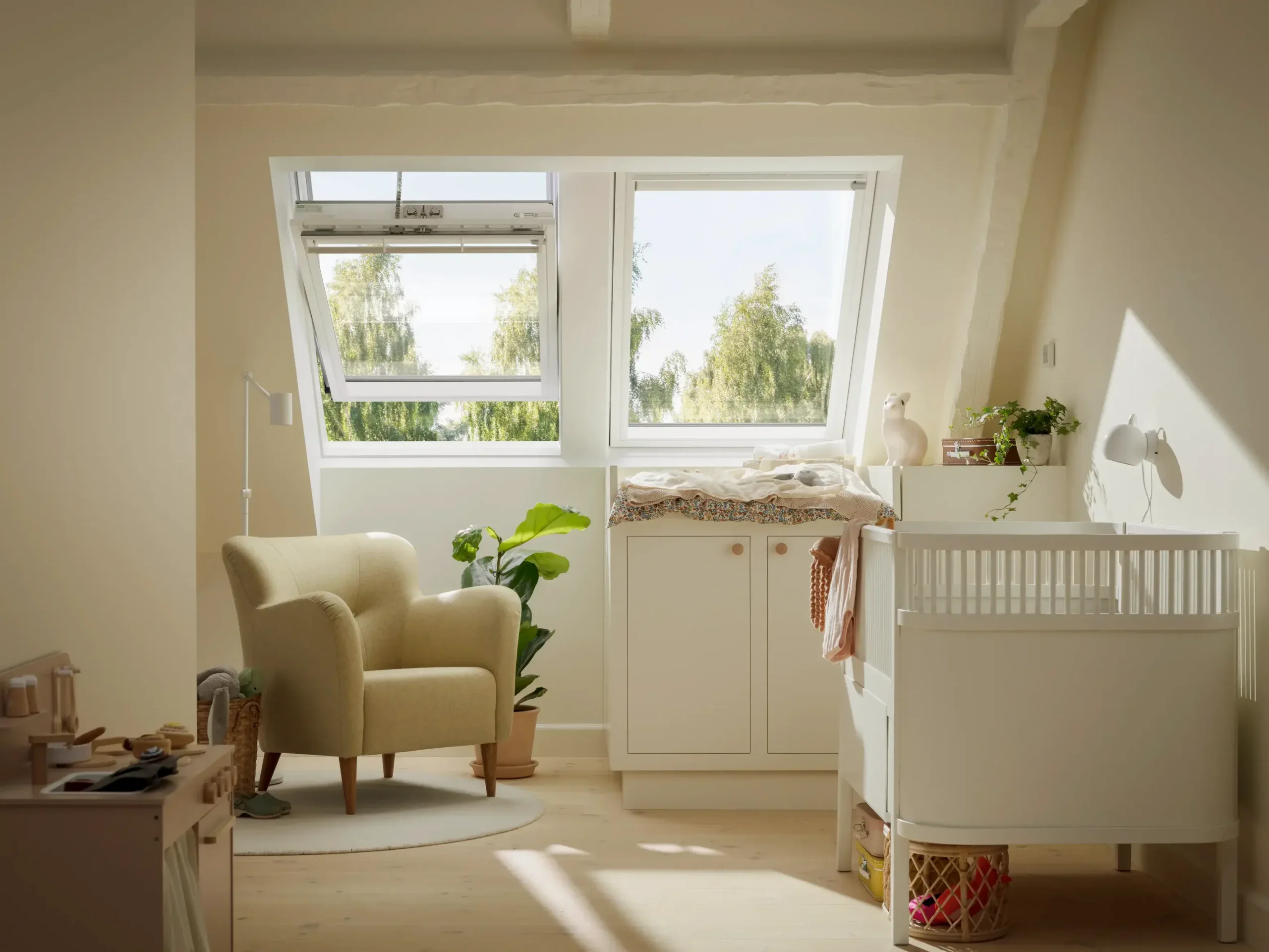 Lyse hvidt børneværelse med store skrå vinduer, bløde møbler og planter.